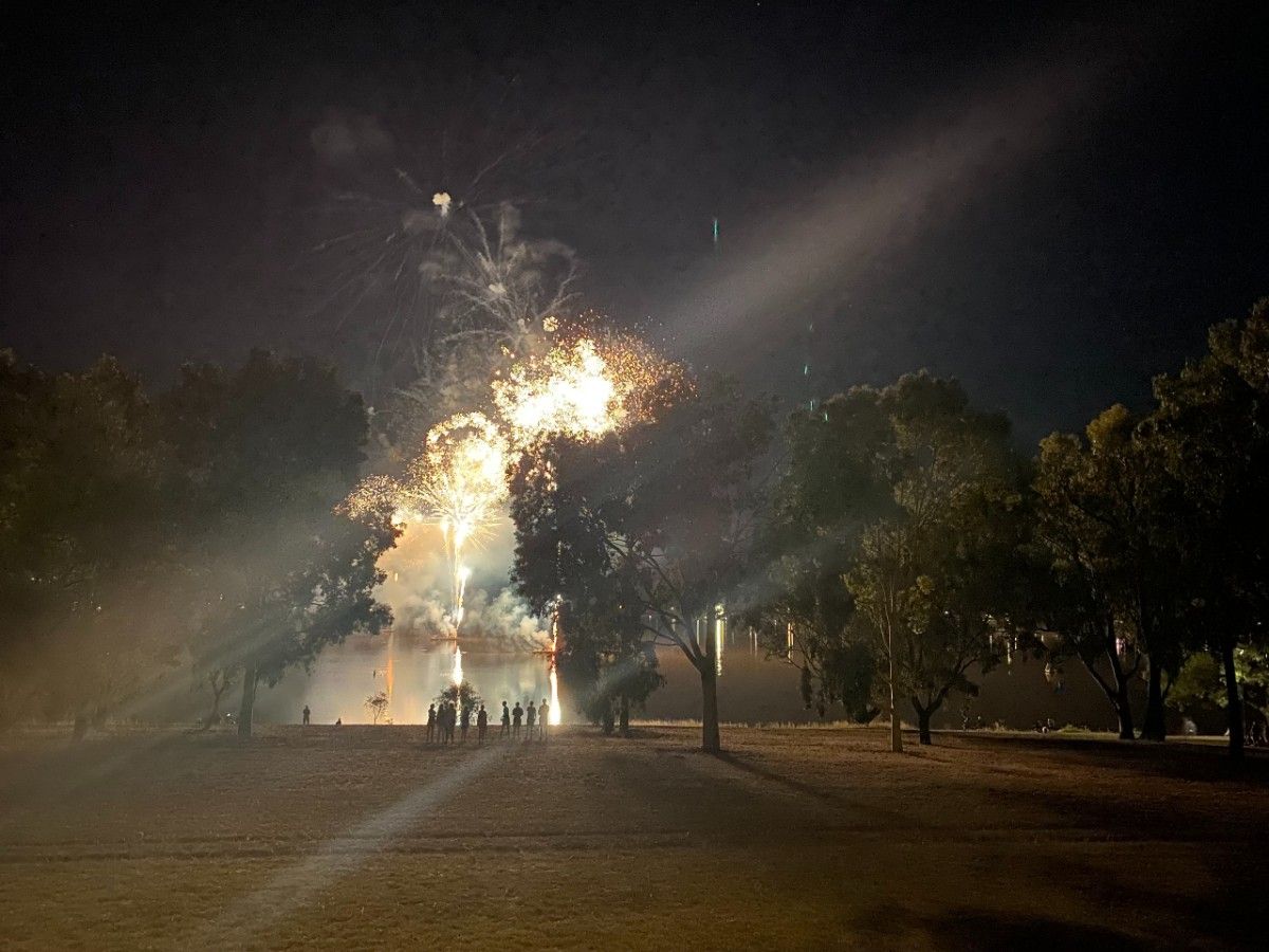 Fireworks explode above a lake with people silhouetted in the foreground