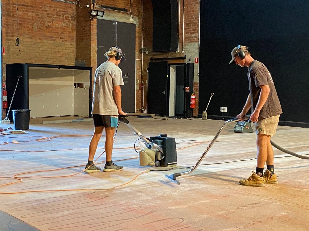 Two men working on a stage surface, one with a sander, the other with a vacuum cleaner.