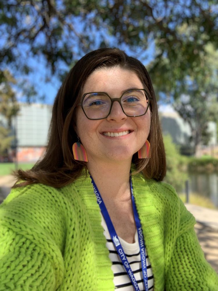 A portrait photograph of a young woman with light brown hair, black rim glasses, orange dangle earrings and a chunky knit green cardigan. 