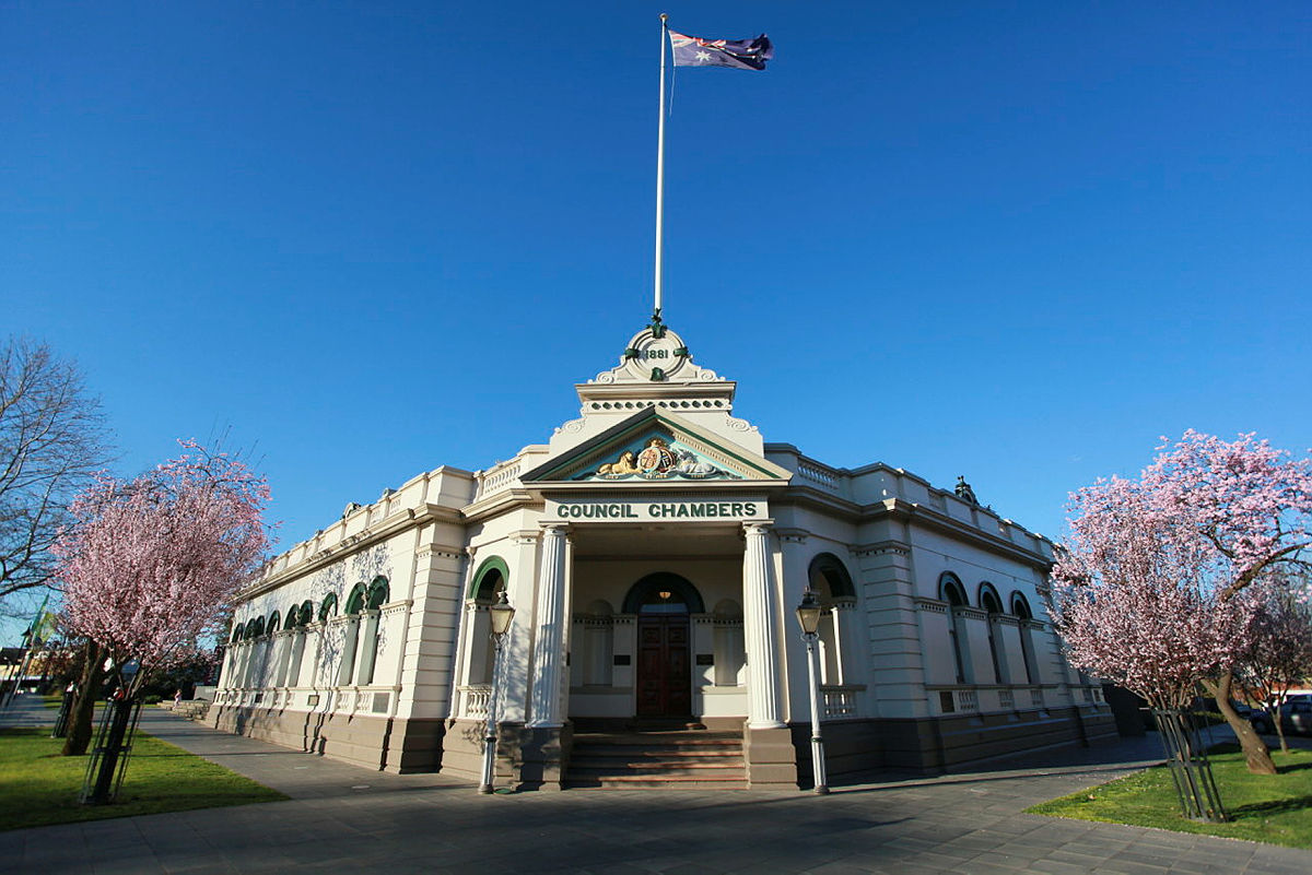 An old administration building on a street corner. On the front of the building are the words "Council Chambers". 