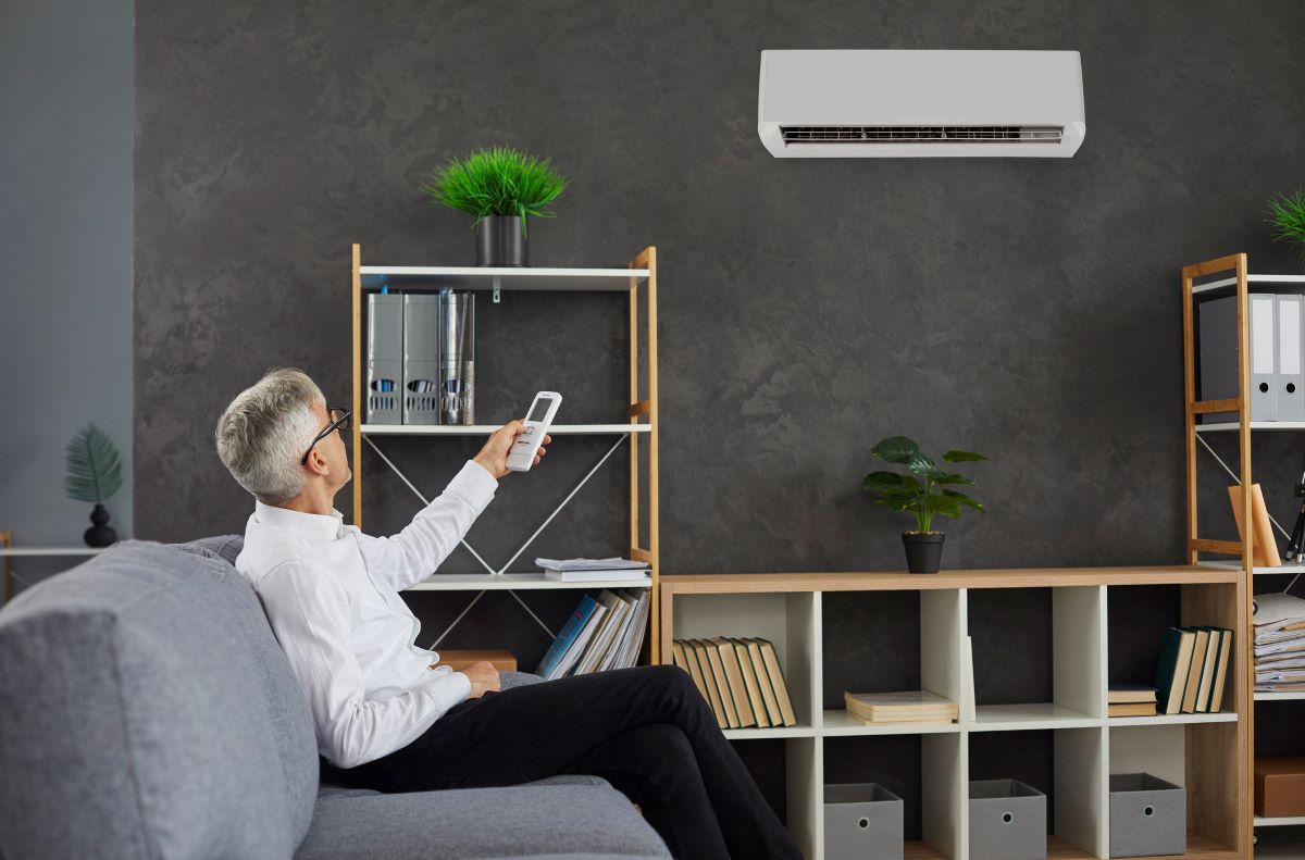 A man with grey hair sitting on a grey lounge turning on a split-system air conditioner mounted on the wall above him. 