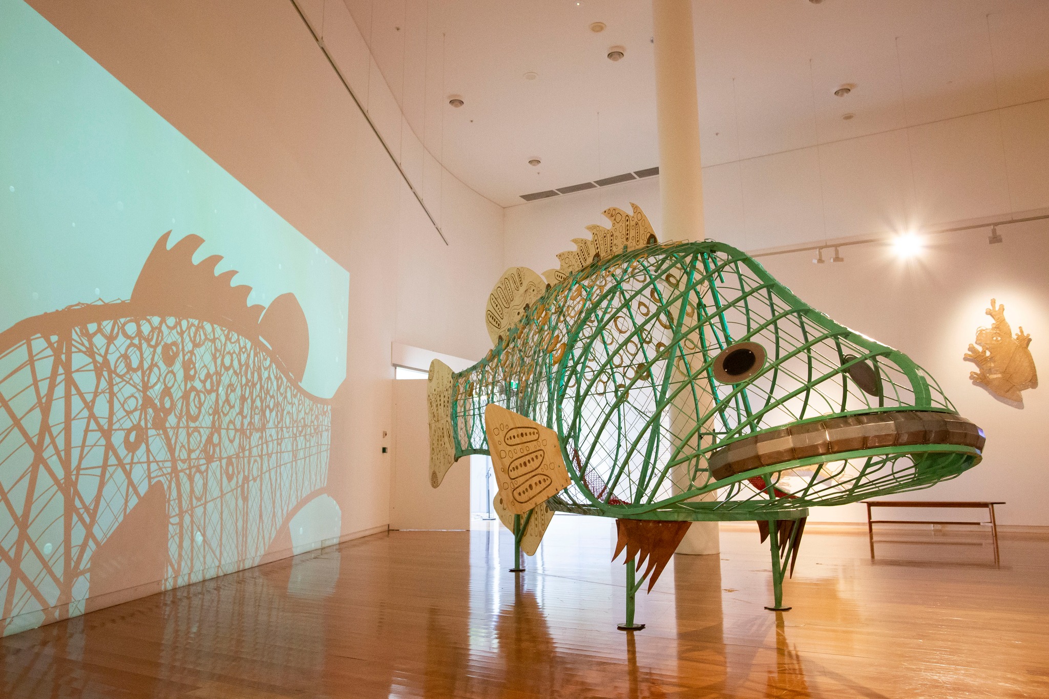 A large sculpture of a trout fish in an art gallery space. There is a projection on the wall to the left. 