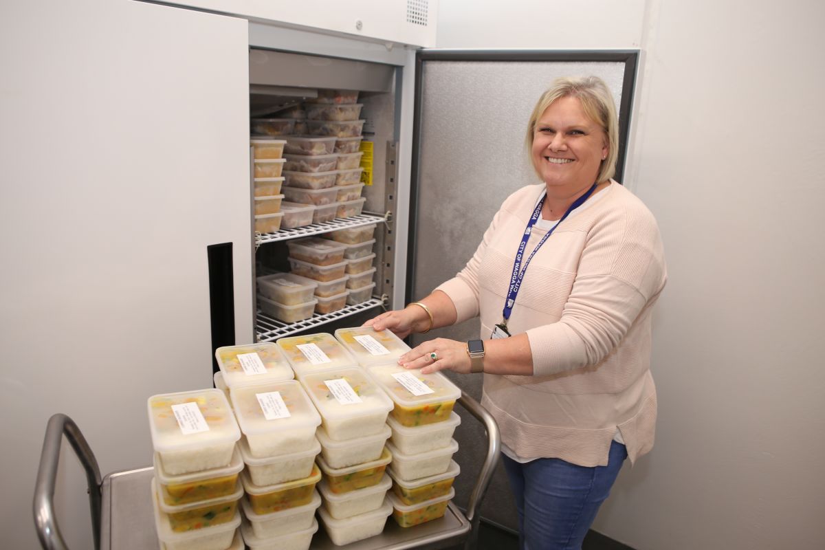 Woman standing next to trolley stacked with pre-prepared meals in plastic cartons, with industrial fridge in background