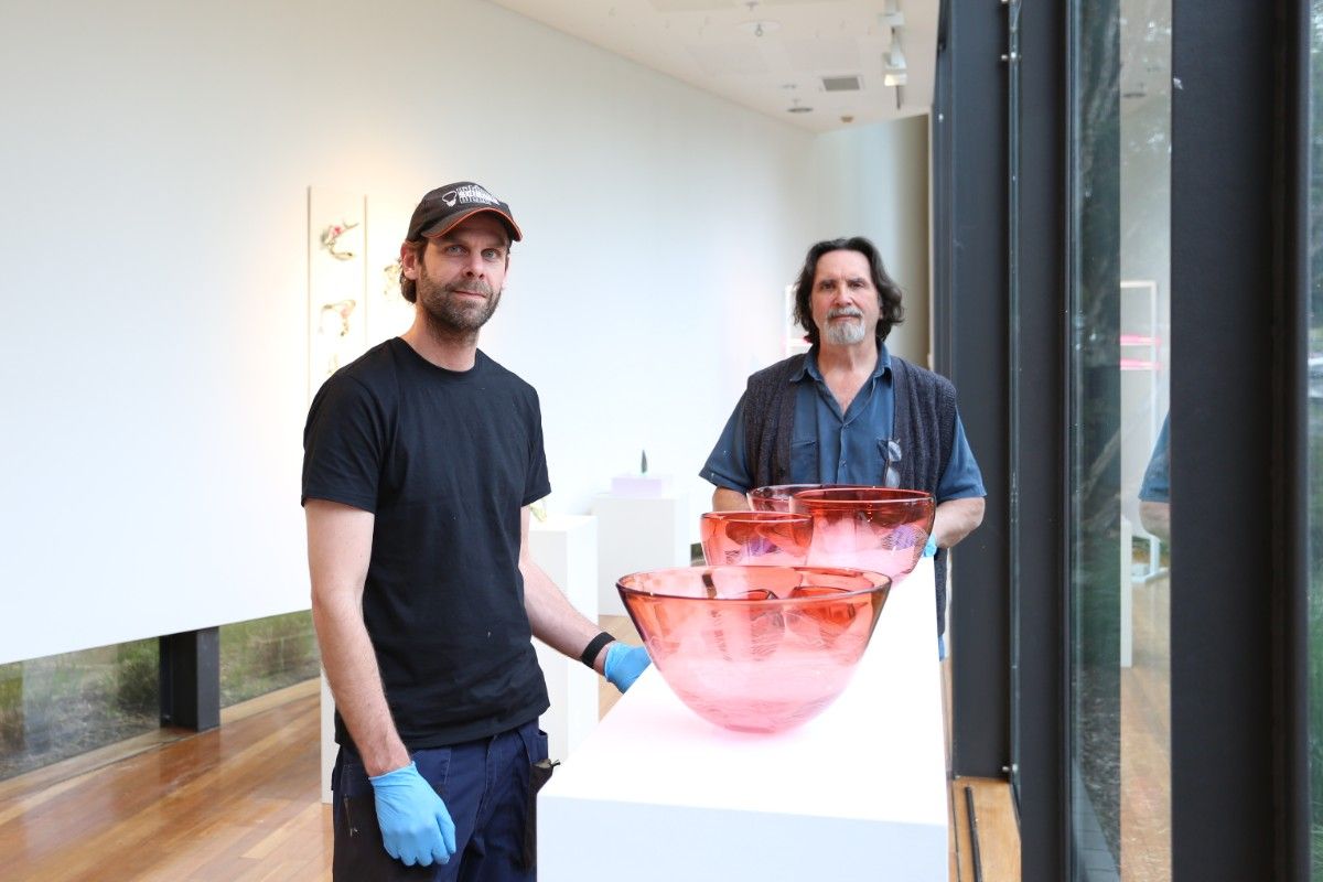Two men wearing blue gloves stand beside a series of large red glass bowls in a gallery setting